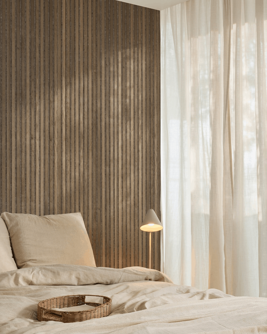 Chambre minimaliste avec linge de lit beige, un plateau en osier, un panneau acoustique à lattes WoodUpp Akupanel Rustic Grey Oak, une lampe blanche et des rideaux voilés.