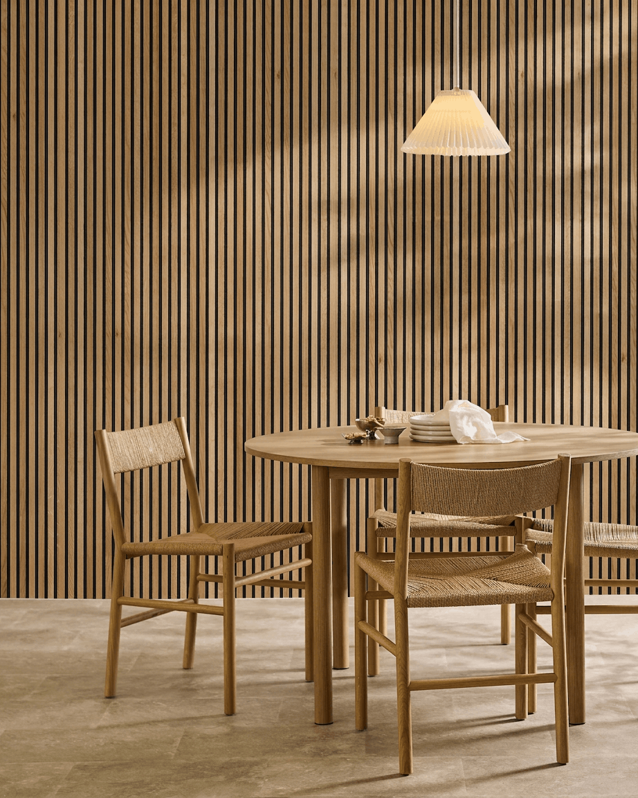 Salle à manger minimaliste avec une table en bois, deux chaises et un panneau acoustique en bois WoodUpp Akupanel en chêne Rustic Brown (300 x 60 cm), éclairée par une suspension.