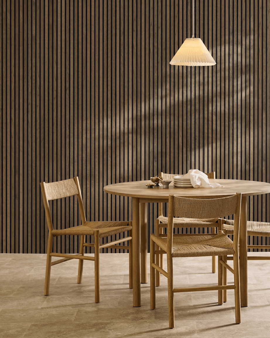 Salle à manger minimaliste avec table ronde, chaises tressées et suspension, mettant en valeur un panneau mural acoustique à lattes WoodUpp Akupanel Rustic Grey Oak.