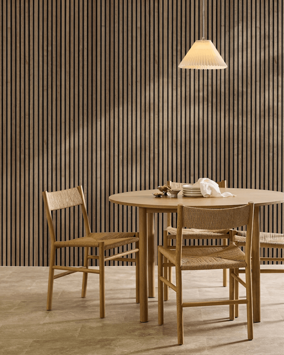 Salle à manger minimaliste avec une table ronde, des chaises tressées, une suspension, et un panneau acoustique en bois WoodUpp Akupanel en Natural Wood avec du feutre black au mur.