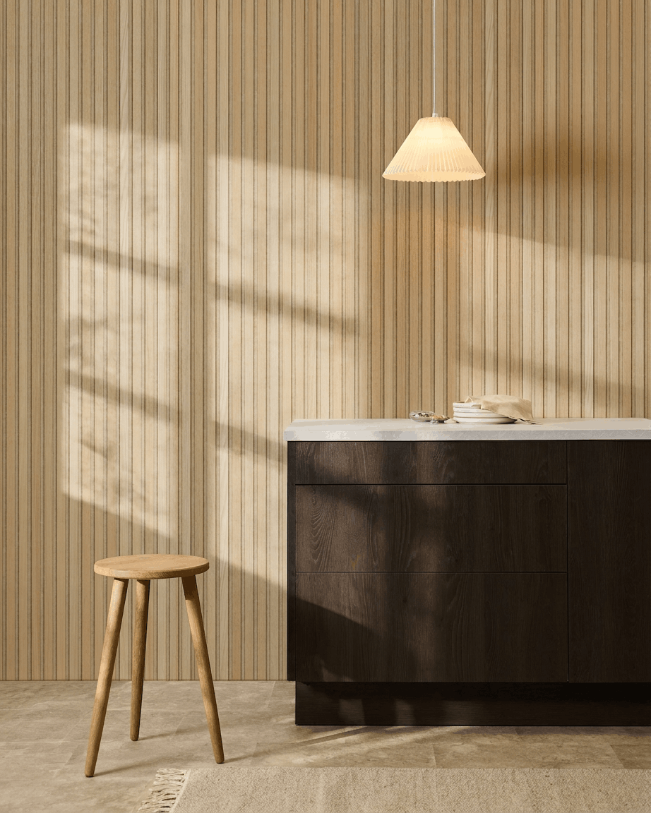 Cuisine scandinave avec le panneau mural acoustique à lattes WoodUpp Akupanel Classic Oak, des meubles en bois foncé, un tabouret en bois, une suspension et la lumière du soleil projetant des ombres.