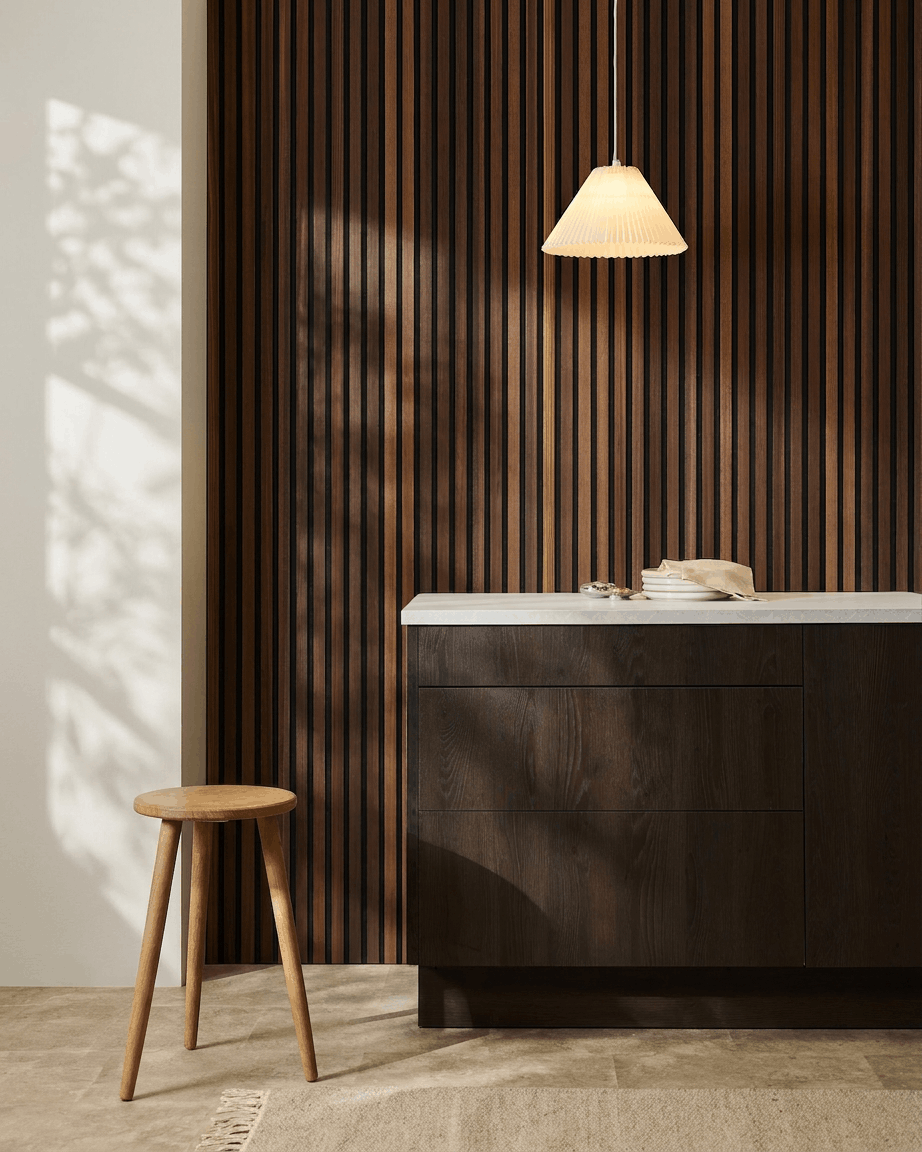 Intérieur minimaliste avec un tabouret en bois, un meuble en bois foncé, une suspension, et un panneau mural acoustique à lattes en bois WoodUpp Akupanel en Natural Wood/mélèze fumé.