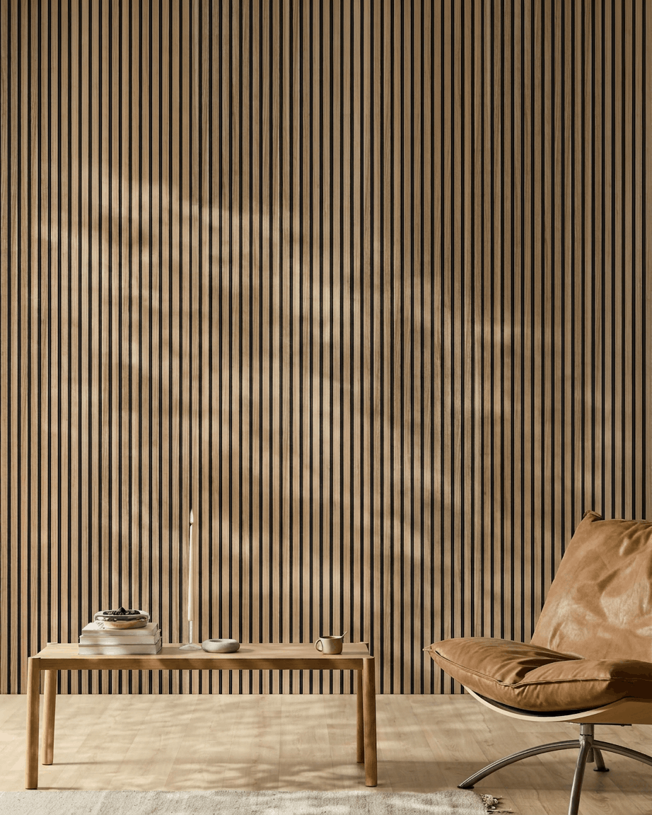 Minimalistischer Raum mit einem WoodUpp Akupanel Akustikpaneel (English-Walnut-Lamellen, black Filz), einem hellbraunen Ledersessel, einem Tisch aus hellem Holz, einer Kerze, Büchern, einer Tasse und einem weißen Teppich im Sonnenlicht.