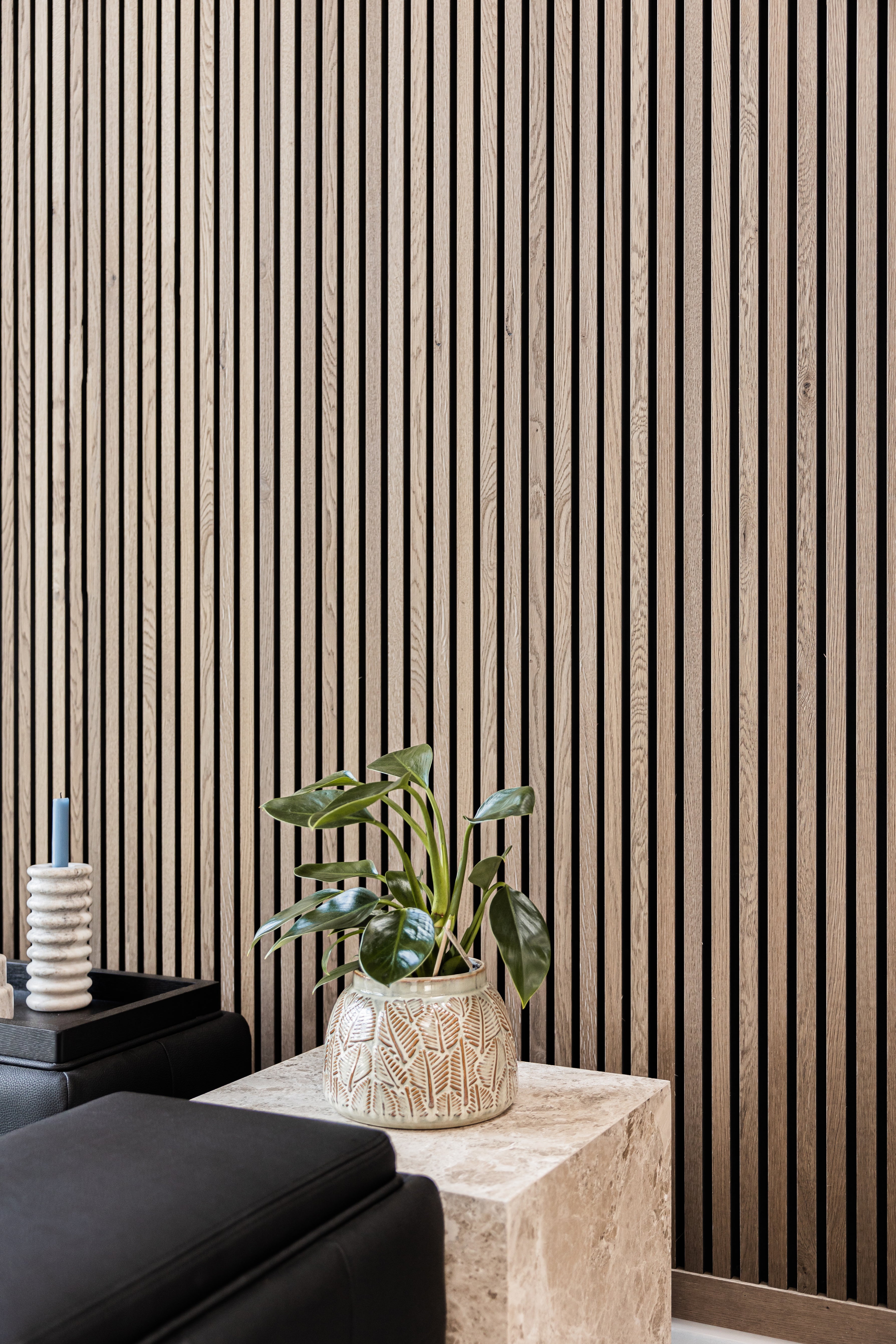 A potted plant sits on a stone table beside black furniture and a candle holder, in front of a warm-toned Akupanel acoustic wood slatwall panel.
