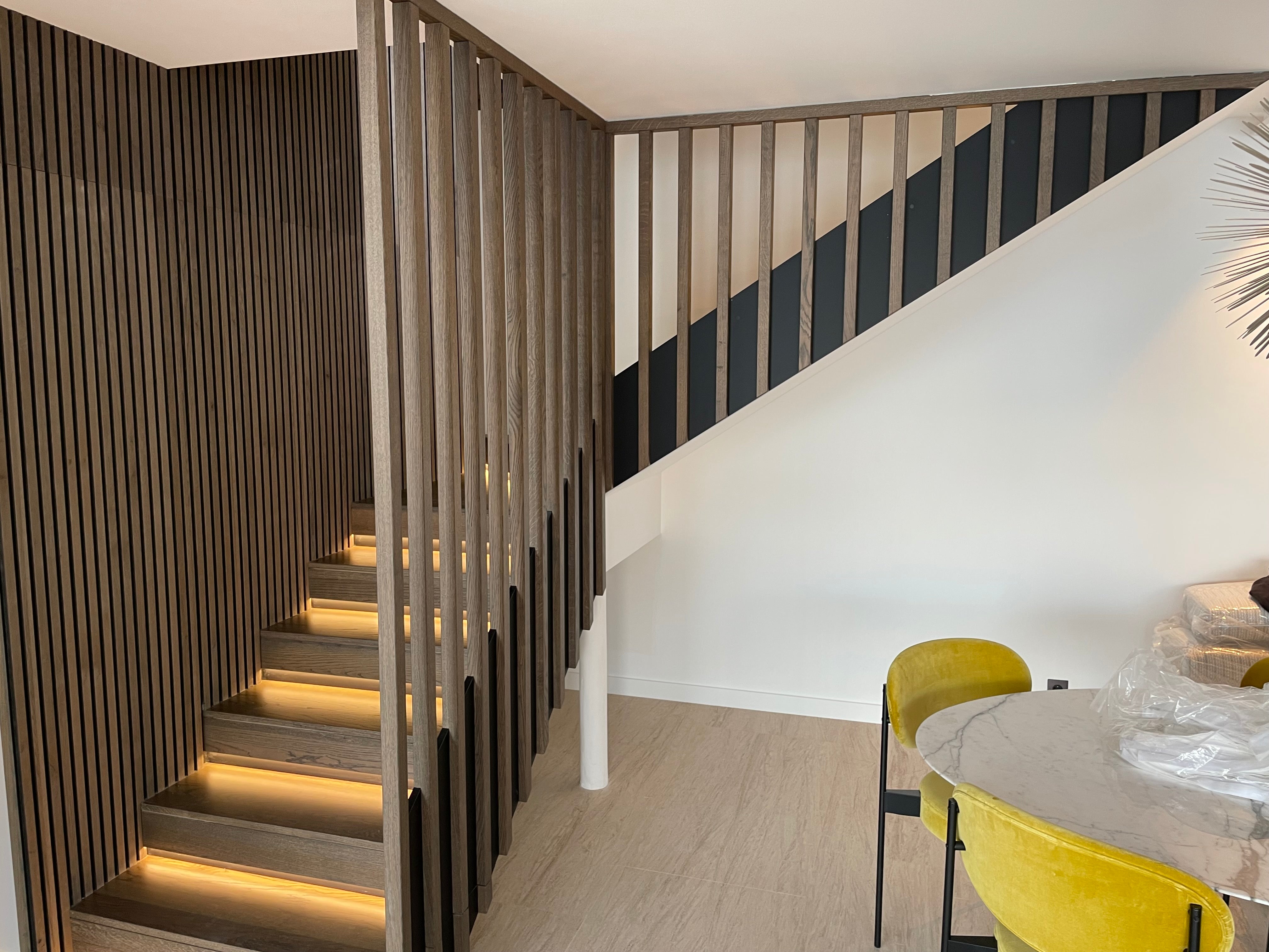 Modern wooden staircase with LED-lit steps, Akupanel vertical acoustic slat panels, yellow velvet chairs, and a round marble table in a Nordic interior.