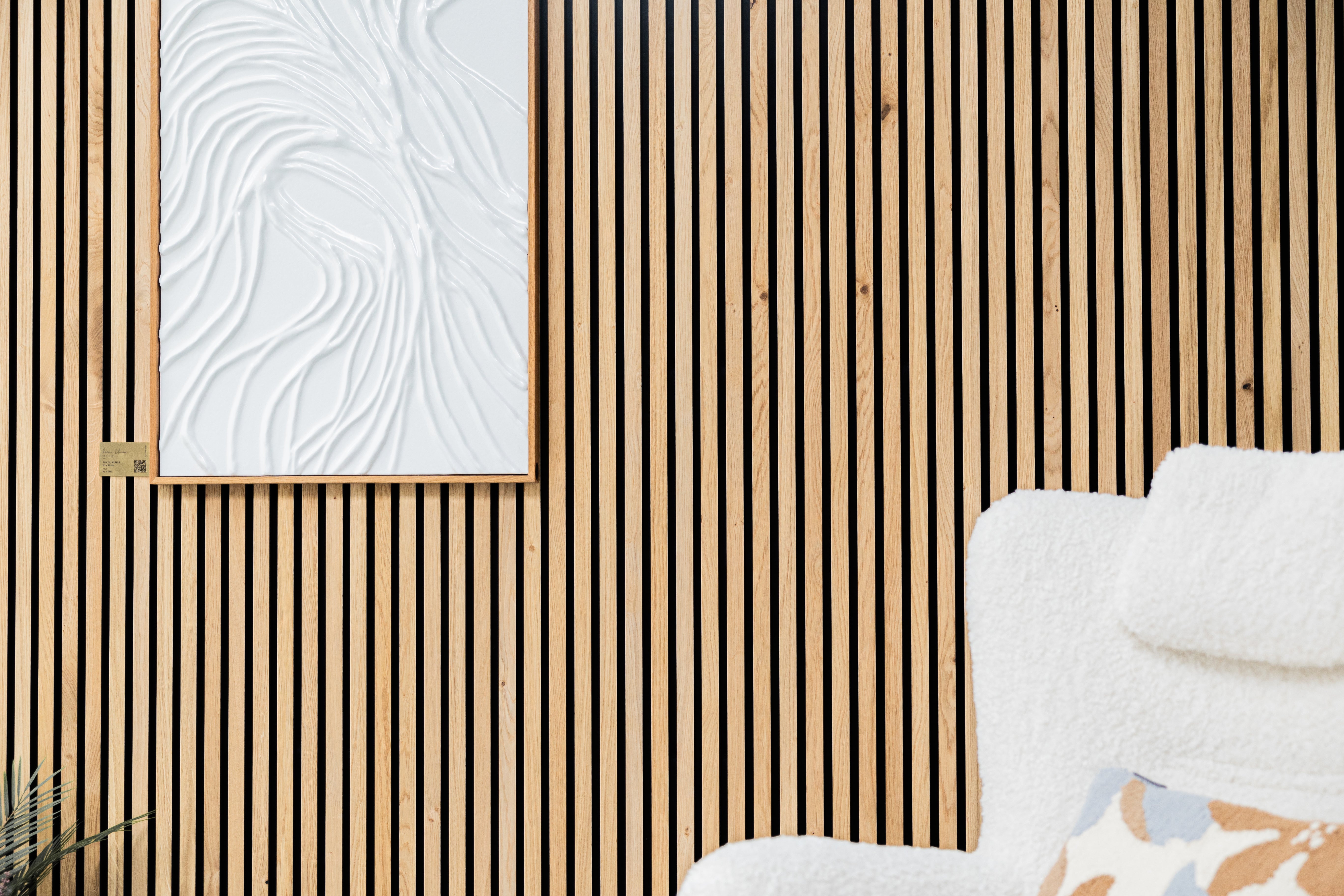 A white textured acoustic Akupanel is mounted on a light wood slat wall behind a fuzzy white chair with a patterned pillow in the lower right corner.