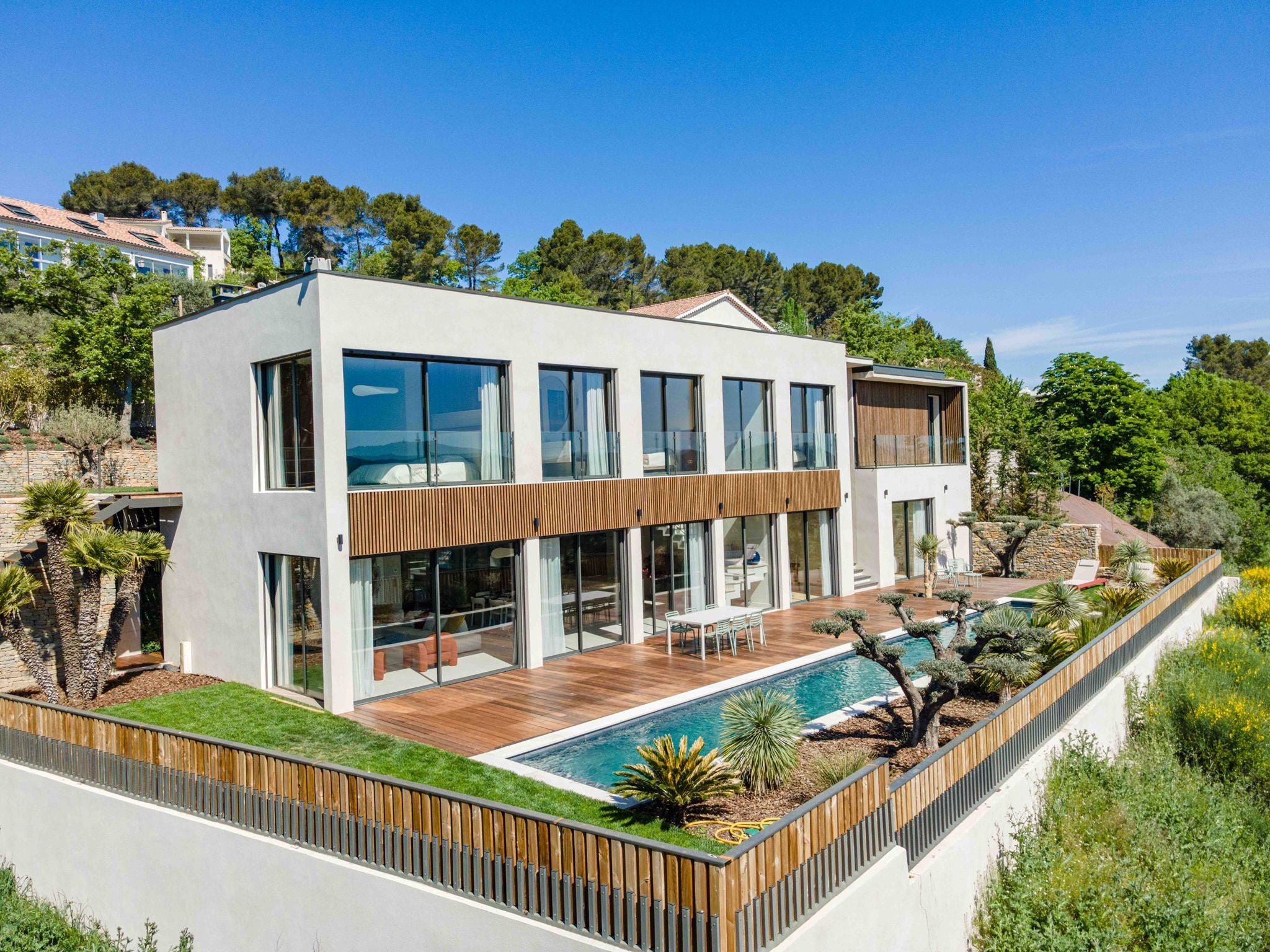 Modernes zweistöckiges Haus mit großen Fenstern, nordischen Holzfassadenelementen, einem Pool, einer großzügigen Terrasse und viel Grün unter strahlend blauem Himmel.