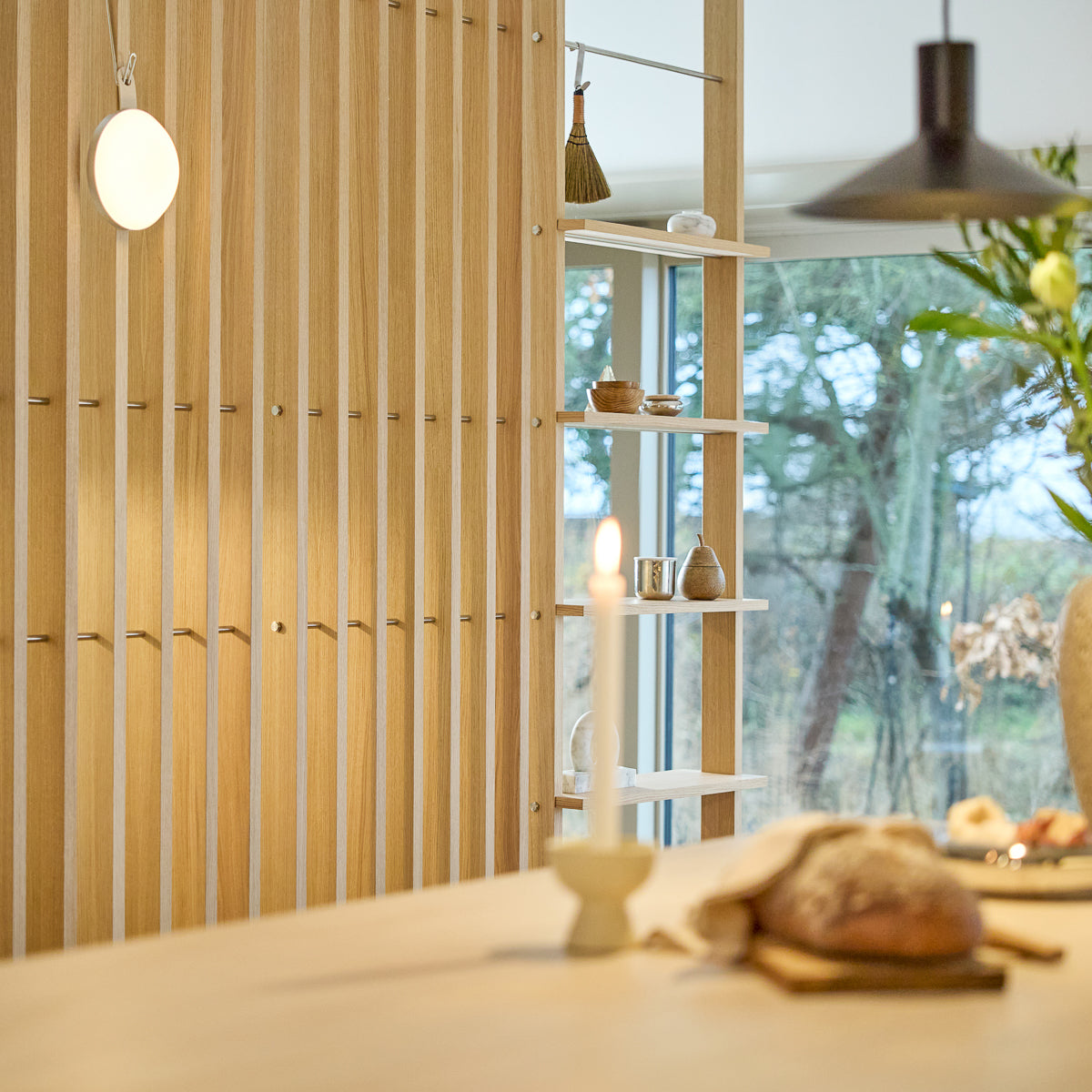 Minimalistisches Interieur mit einem Nordic EMBRACE Raumteiler aus Holz, einer brennenden Kerze, frischem Brot auf dem Tisch, ausgewählter Dekoration auf den Regalen und einem Fenster, das draußen Bäume einrahmt.