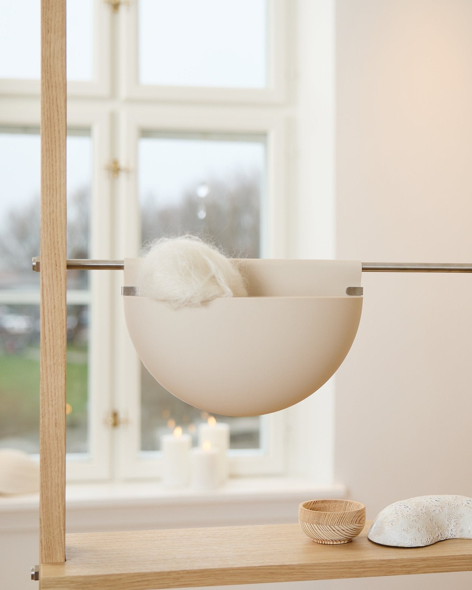 Un bol blanc avec un objet duveteux repose sur une étagère en bois, près de bougies, d’objets déco et d’un accessoire acoustique WoodUpp Gro Soft Pocket BM2 en beige clair, près de la fenêtre.