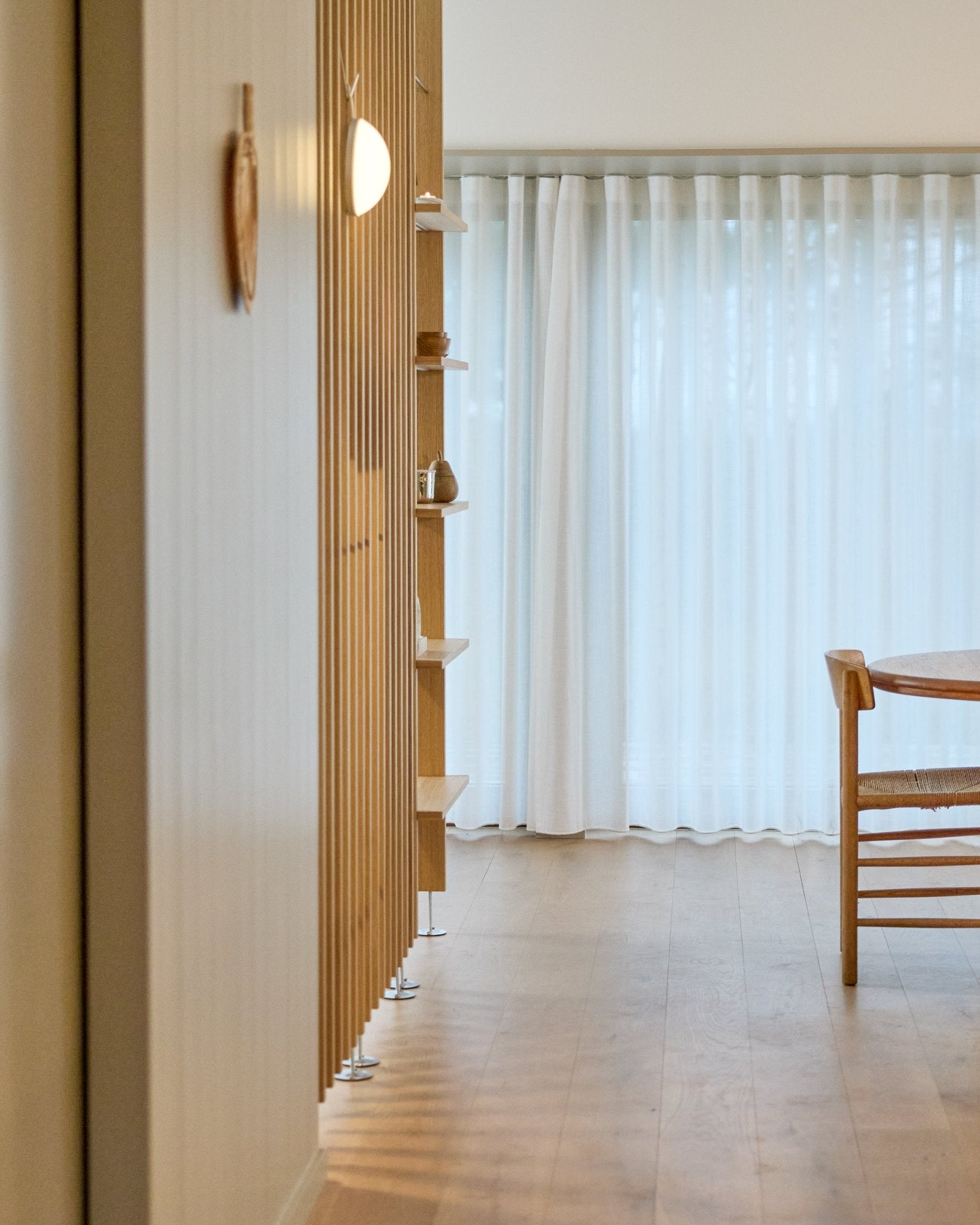 Pièce minimaliste avec une cloison de séparation WoodUpp Embrace du sol au plafond, un sol en bois, des étagères, une chaise et des rideaux voilés devant de large fenêtres.