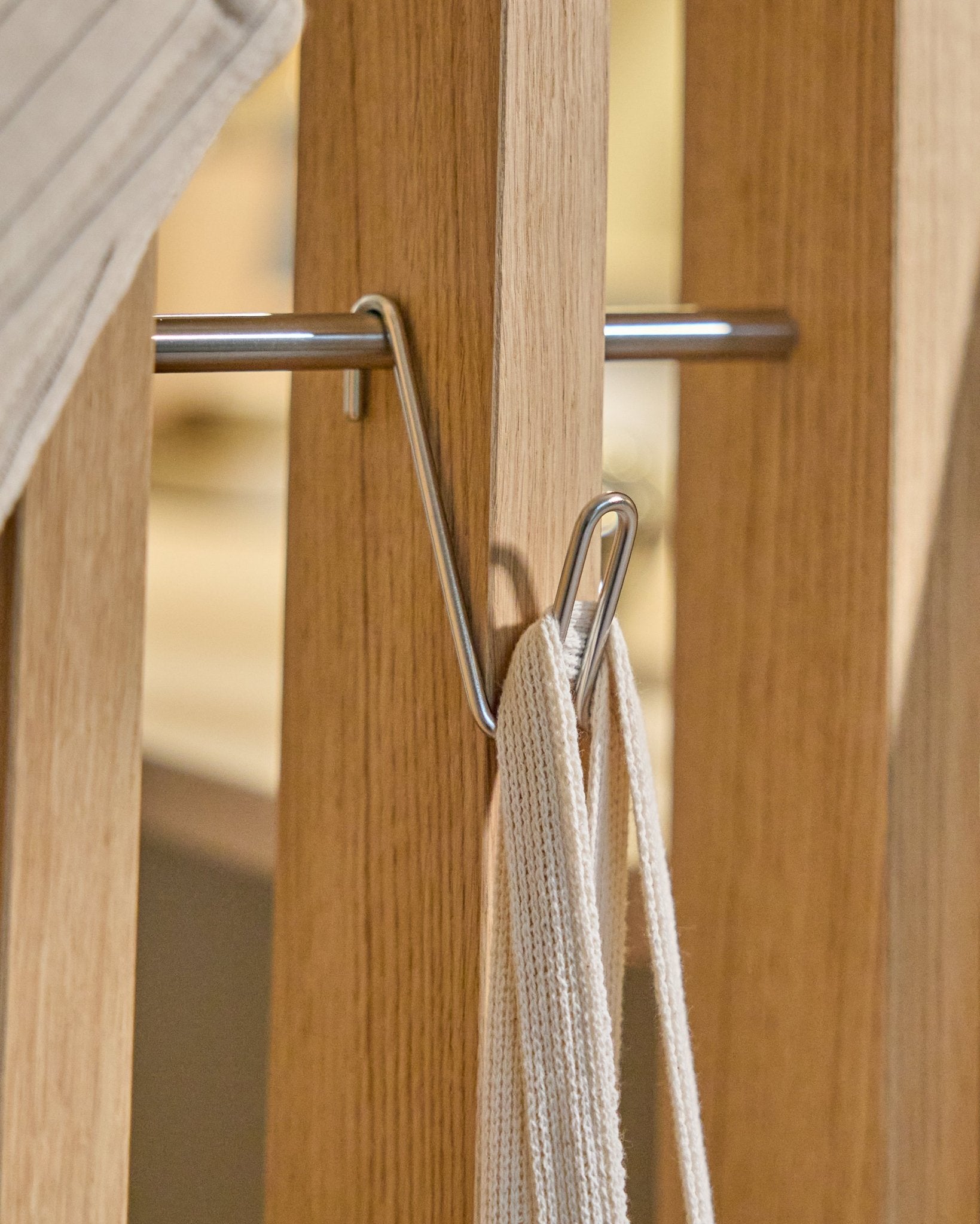 La patère filaire Alva / BM1 (Stainless Steel, Short) de WoodUpp est montée sur une latte en bois de Panneau acoustique et maintient une sangle blanche avec tige métallique ; fond beige.