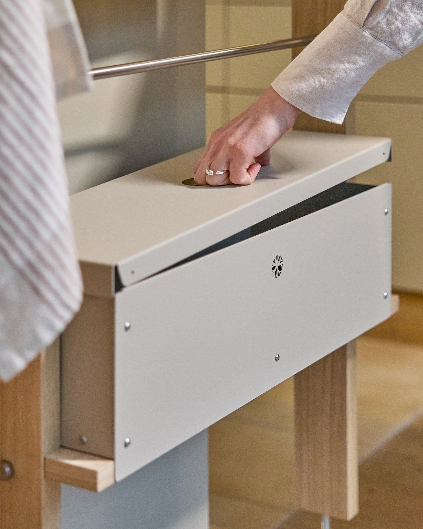 Une main soulève le couvercle du WoodUpp Nell Metal Box BM2 en beige, avec structure en steel thermolaqué et supports en bois, conçu pour un rangement nordique bien organisé.