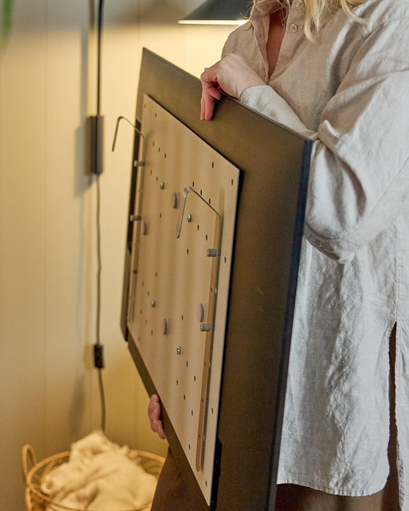 Une personne en chemise claire tient le WoodUpp Nell BM1 TV-Mount en beige, 64x44 cm — compatible avec les panneaux acoustiques en bois nordiques, pour des installations sans mur.