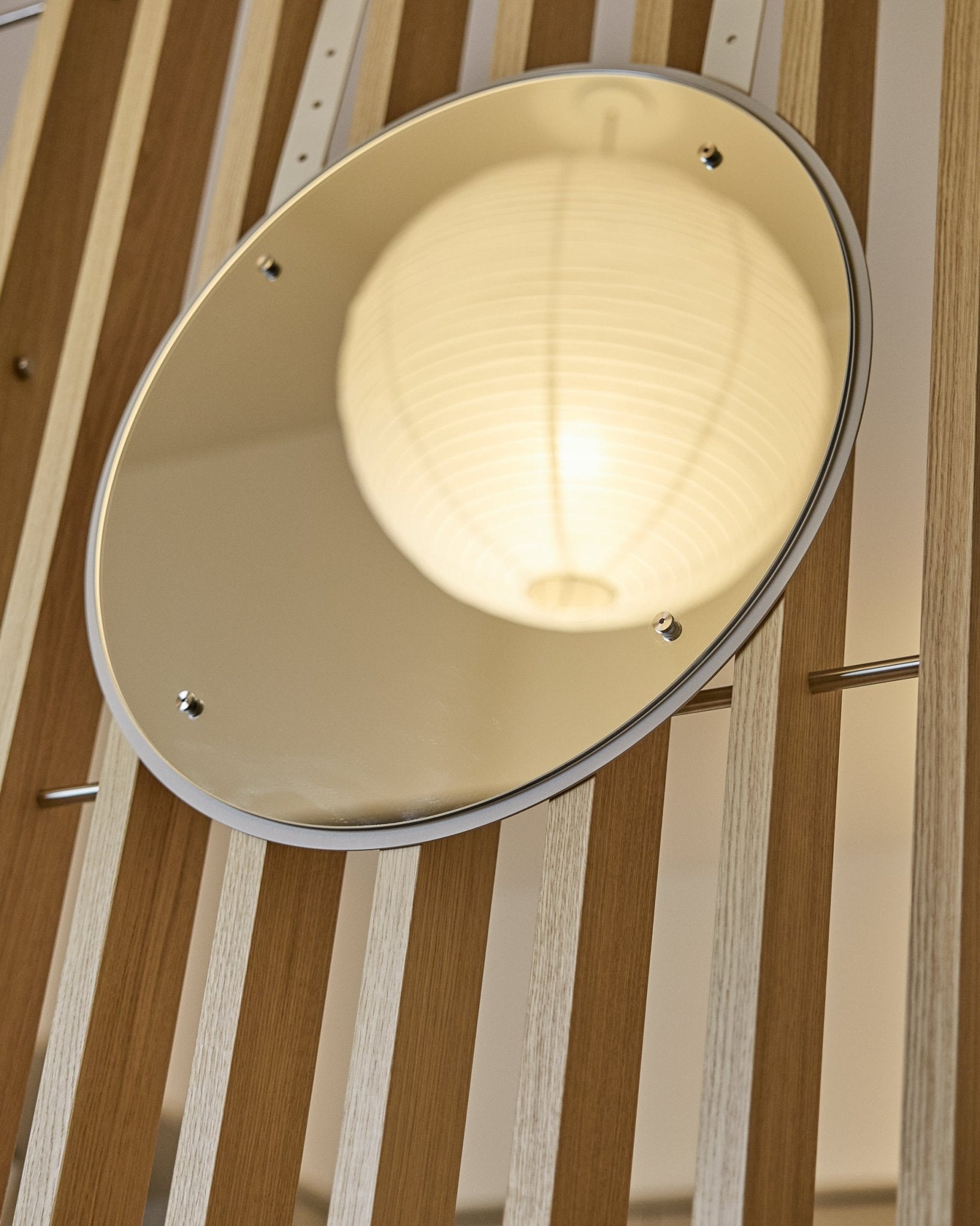 Le Vala | Mirror / BM1, D54 cm de WoodUpp est un miroir rond réglable en hauteur, monté sur des panneaux acoustiques à lattes verticales, qui reflète une lanterne en papier lumineuse.