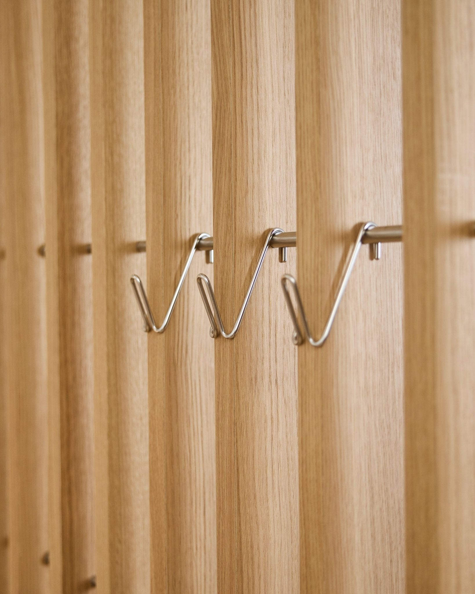 Trois accessoires WoodUpp Alva Wire Hook / BM1 courts en acier inoxydable sont montés à intervalles réguliers sur une rangée sur un panneau acoustique à lattes de bois vertical.