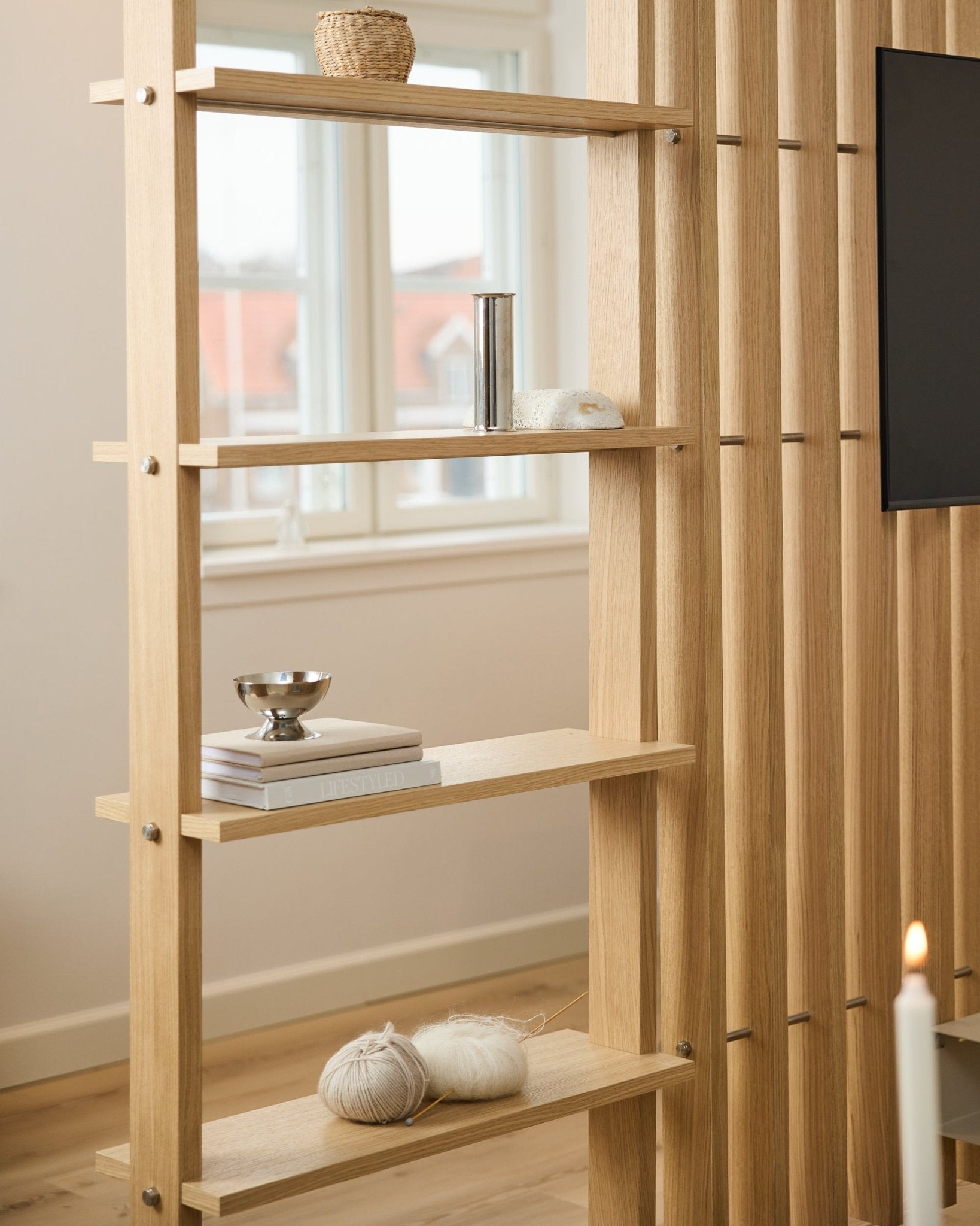 Séparateur de pièce en chêne clair par WoodUpp, avec des lattes verticales et des étagères accueillant des livres et des pelotes de laine.