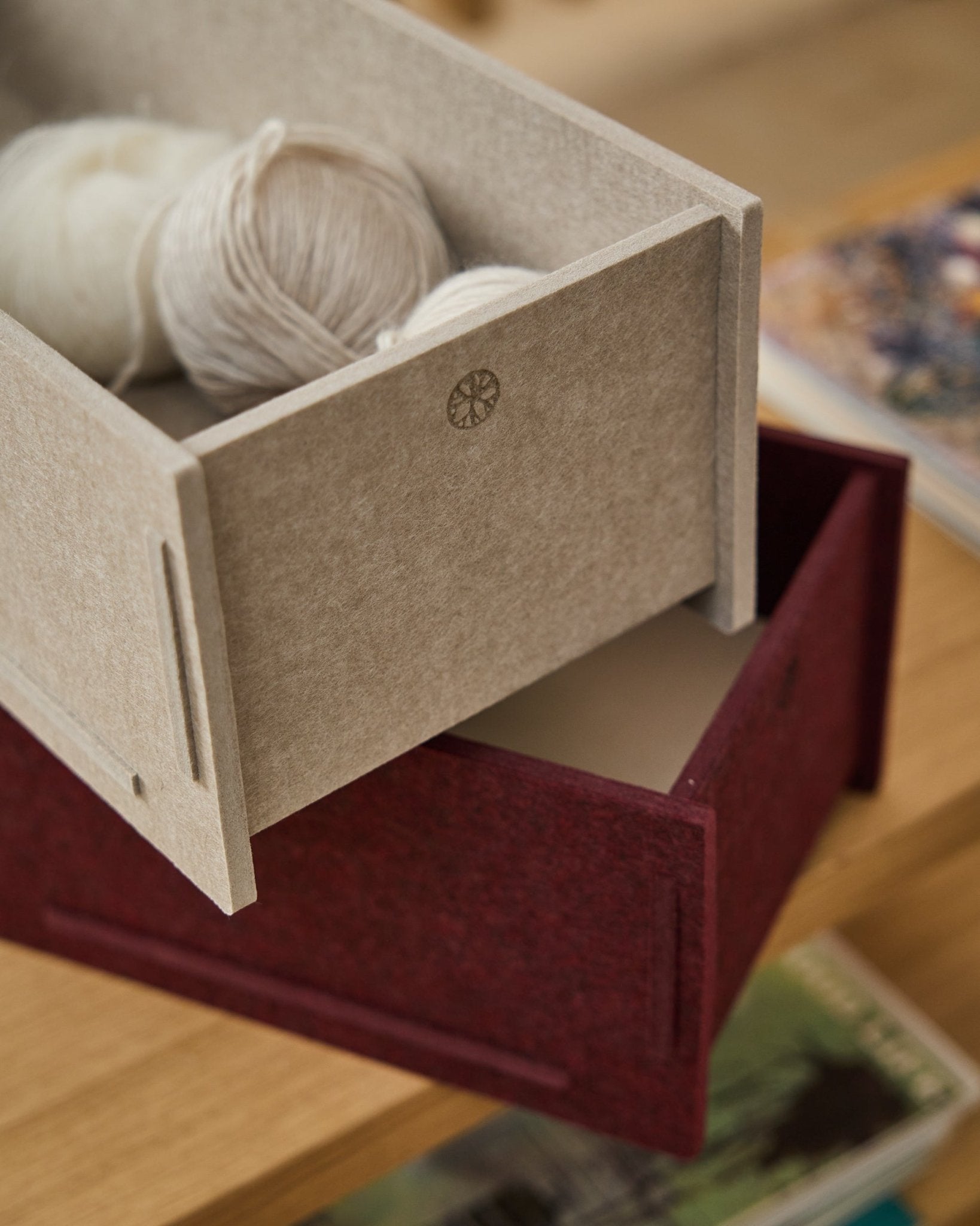 Une boîte de rangement en feutre PET beige remplie de laine repose sur une WoodUpp Vigg Felt Box/BM2 fermée en ox red, posée sur un sol en bois avec des livres en arrière-plan.