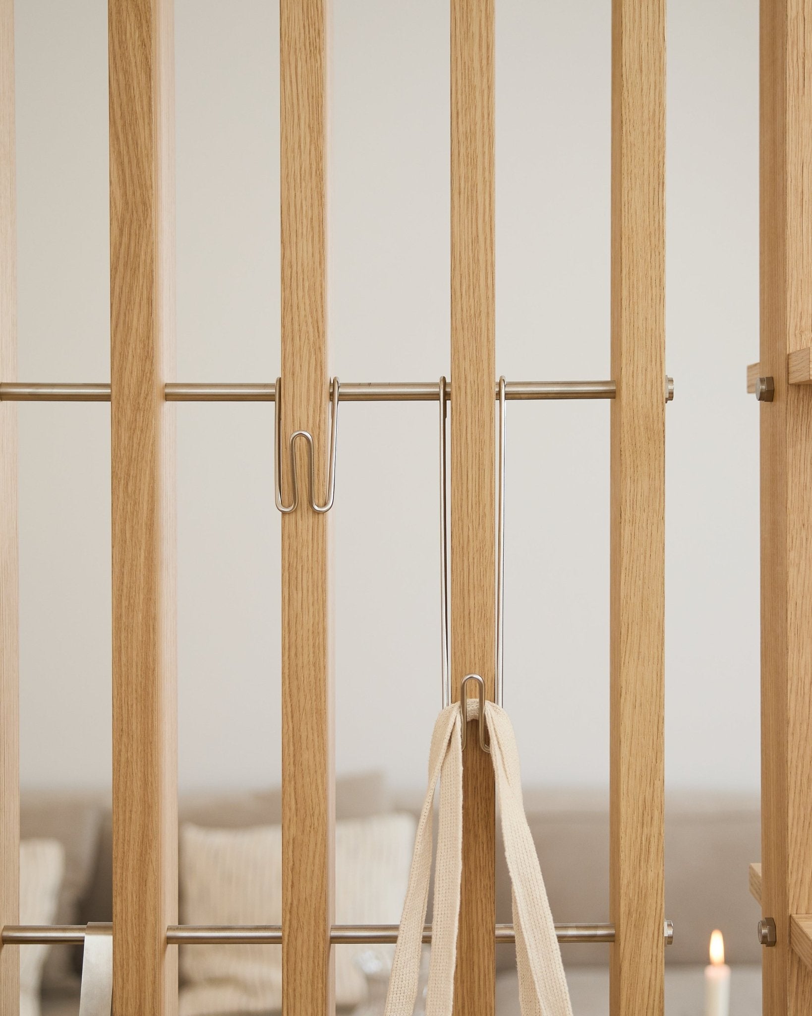 Gros plan sur le WoodUpp Alva | Wire Hook BM1 (acier inoxydable, long, lot de 3) fixé aux lattes d’un Panneau acoustique clair avec une sangle blanche ; canapé et bougie flous en arrière-plan.