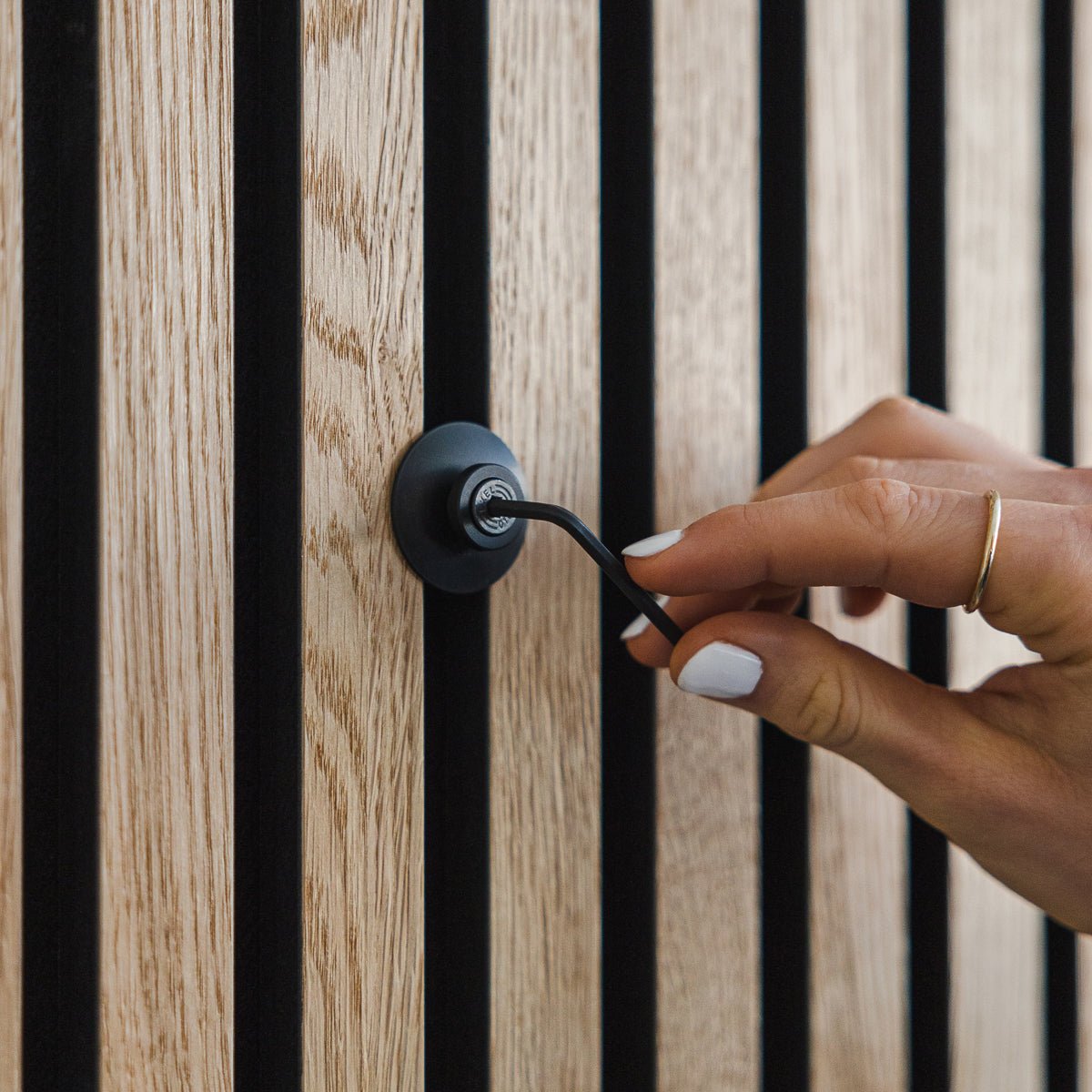 Eine Hand mit weißem Nagellack dreht den Dupps | Heavy Haken aus black Aluminium von WoodUpp an einem verticalen Akupanel Akustik-Holzpaneel.